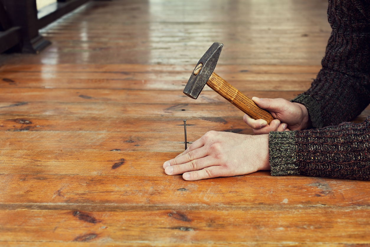 Man driving a nail into an old floor board to stop squeaking
